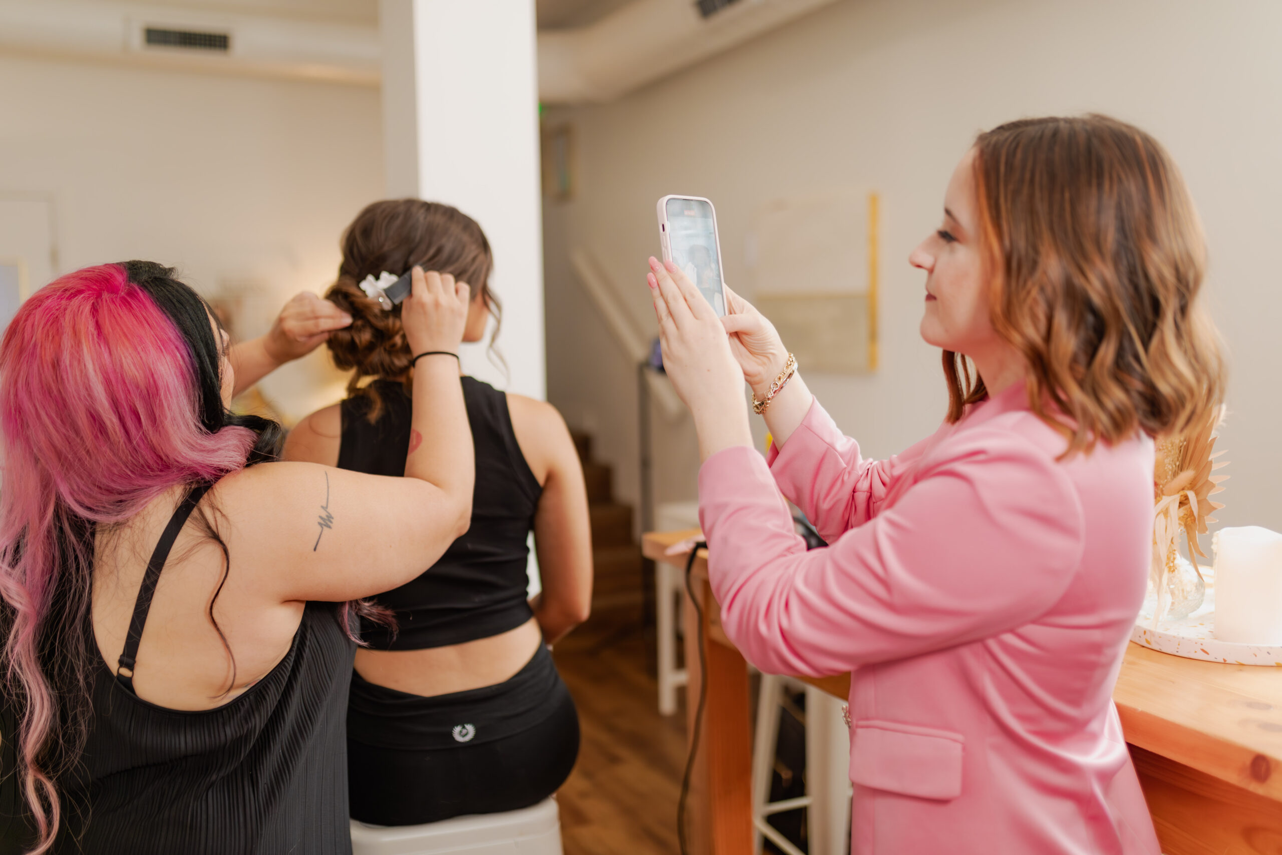 A hairstylist working on an updo while a woman in a pink blazer captures behind-the-scenes content with her phone.